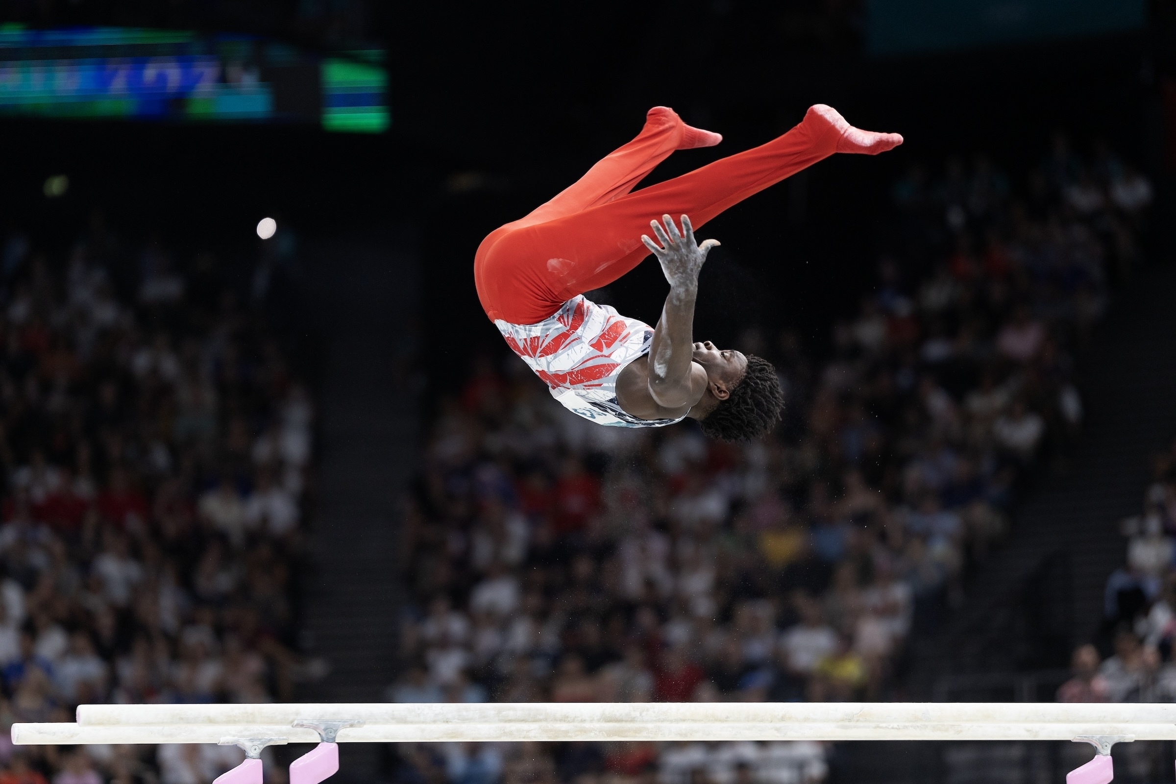 A male gymnast in mid-air performing a backflip during a competition, wearing a red and white unitard