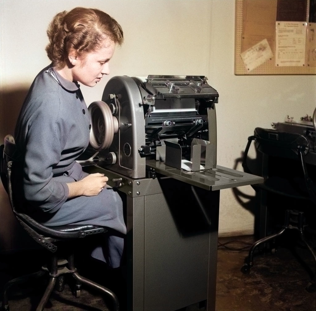 A woman in 1950s attire operates an early office machine, likely a mimeograph or duplicating machine, in a vintage office setting