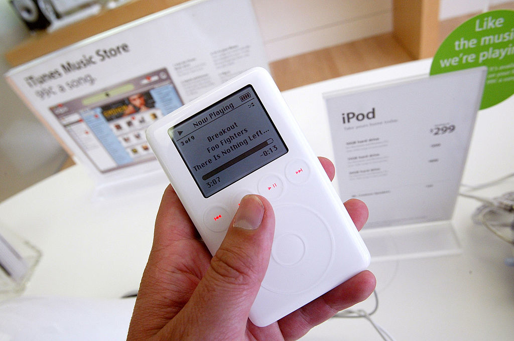 A close-up of a hand holding a classic iPod on display at an Apple Store, showing the song "Breakout" by Foo Fighters playing on the screen. Signs in the background advertise the iPod at $299 and the iTunes Music Store
