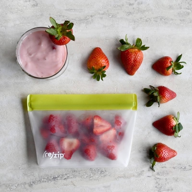 Rezip reusable bag filled with strawberries next to fresh strawberries and a glass bowl of strawberry yogurt
