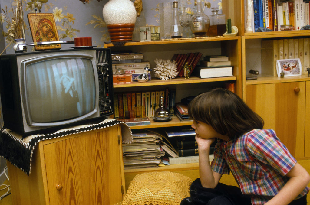 A child watches an old TV in a cozy, book-filled living room, showing a black and white program