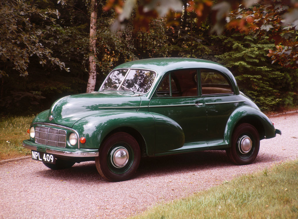 A classic green car, likely a Morris Minor, is parked on a road surrounded by greenery. The license plate reads "NPL 409."