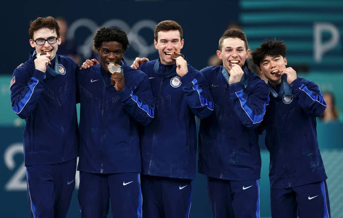 Group of five male athletes in tracksuits, each biting a medal and standing together, celebrating
