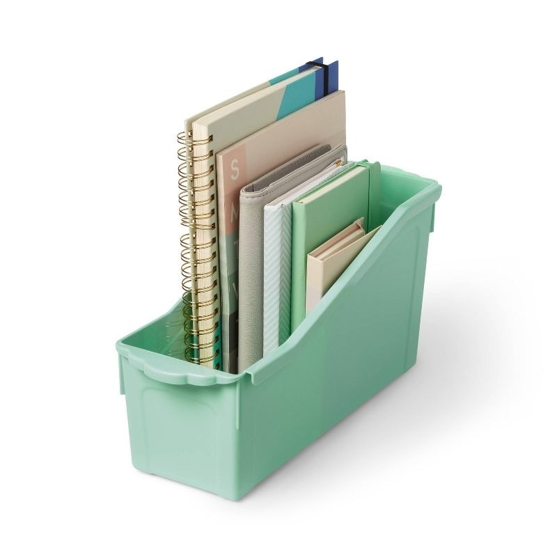 A green plastic storage bin holds various notebooks and folders, organized upright