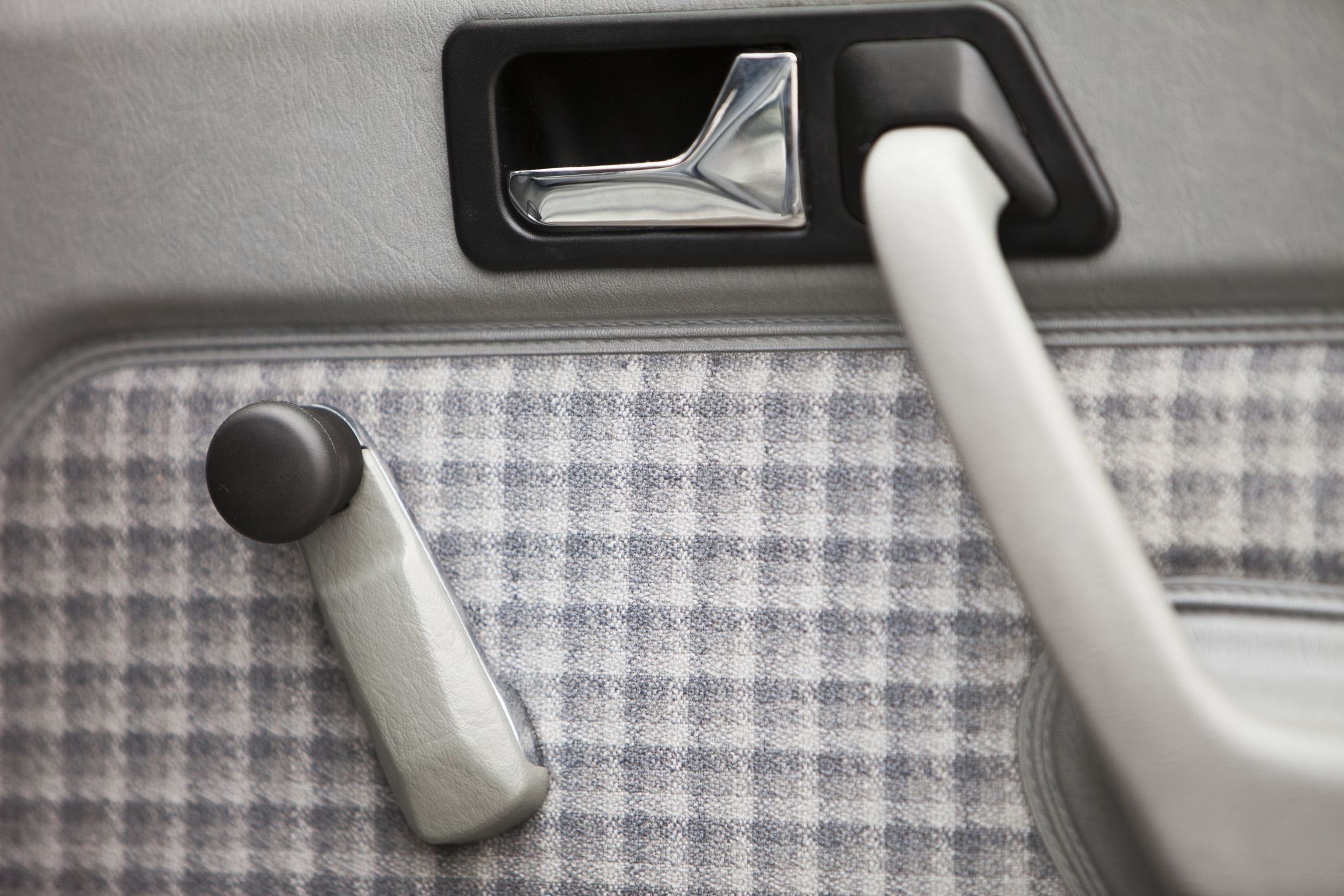 A close-up shot of a car door interior featuring a manual window crank handle and door handle. The panel has a checkered fabric design