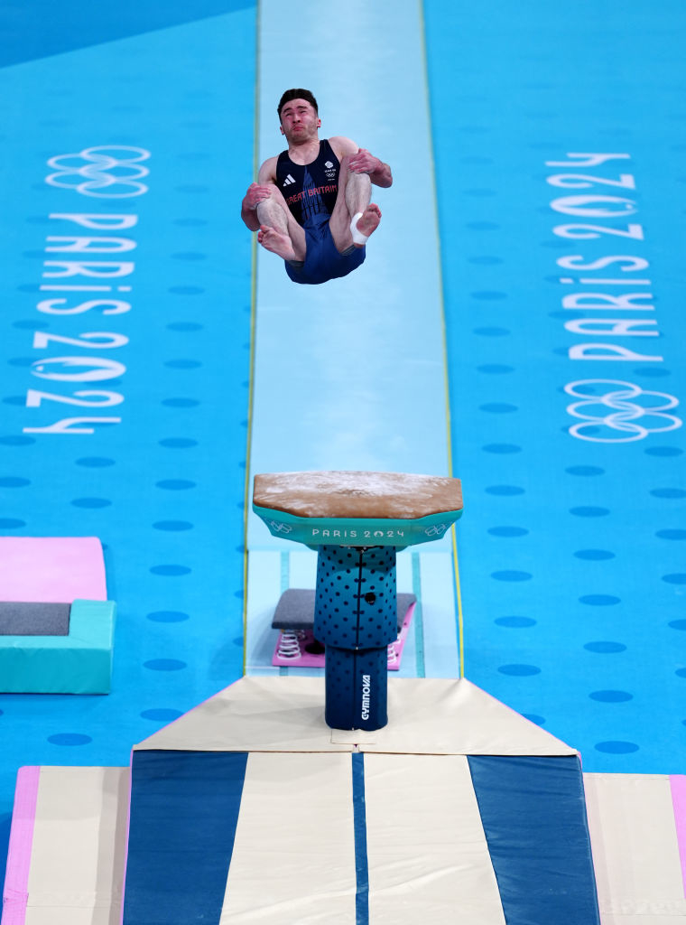 A gymnast performs a high tuck jump on the vault at the 2024 Paris Olympics. The venue features Paris 2024 branding on the floor