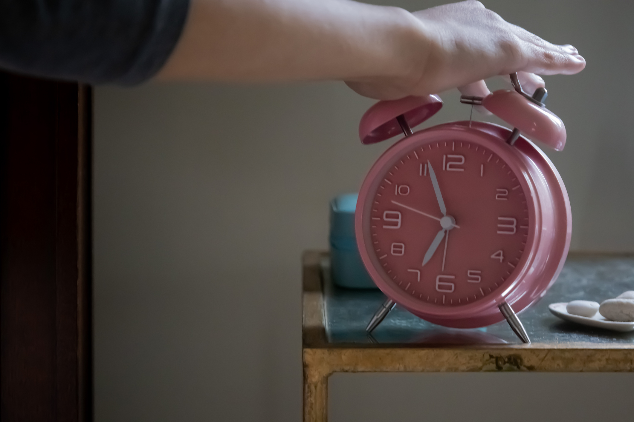 A hand reaches to turn off a ringing pink alarm clock on a small table with two white stones and a blue container