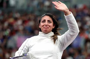 Person in fencing gear waves while appearing emotional in front of a blurred crowd