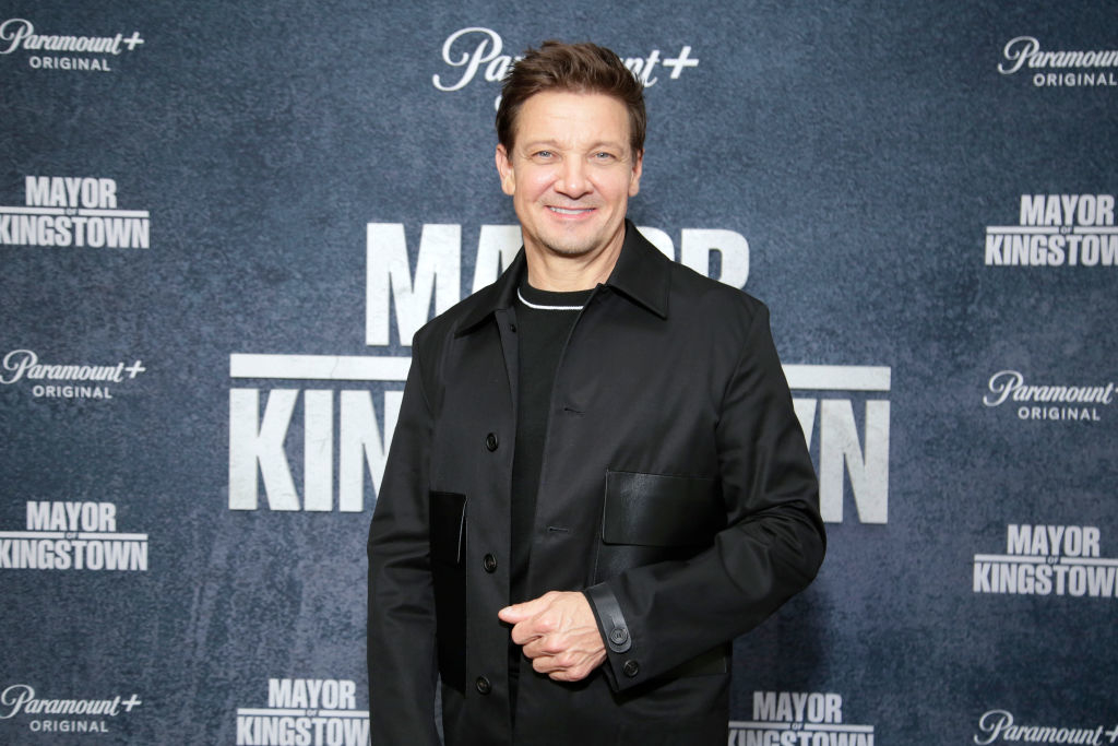 Jeremy Renner at the "Mayor of Kingstown" event, dressed in a button-up coat over a casual shirt, smiling for the camera