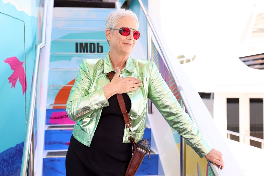 Jamie Lee Curtis wearing a metallic jacket and sunglasses, poses with a hand on her chest near colorful steps with IMDb branding