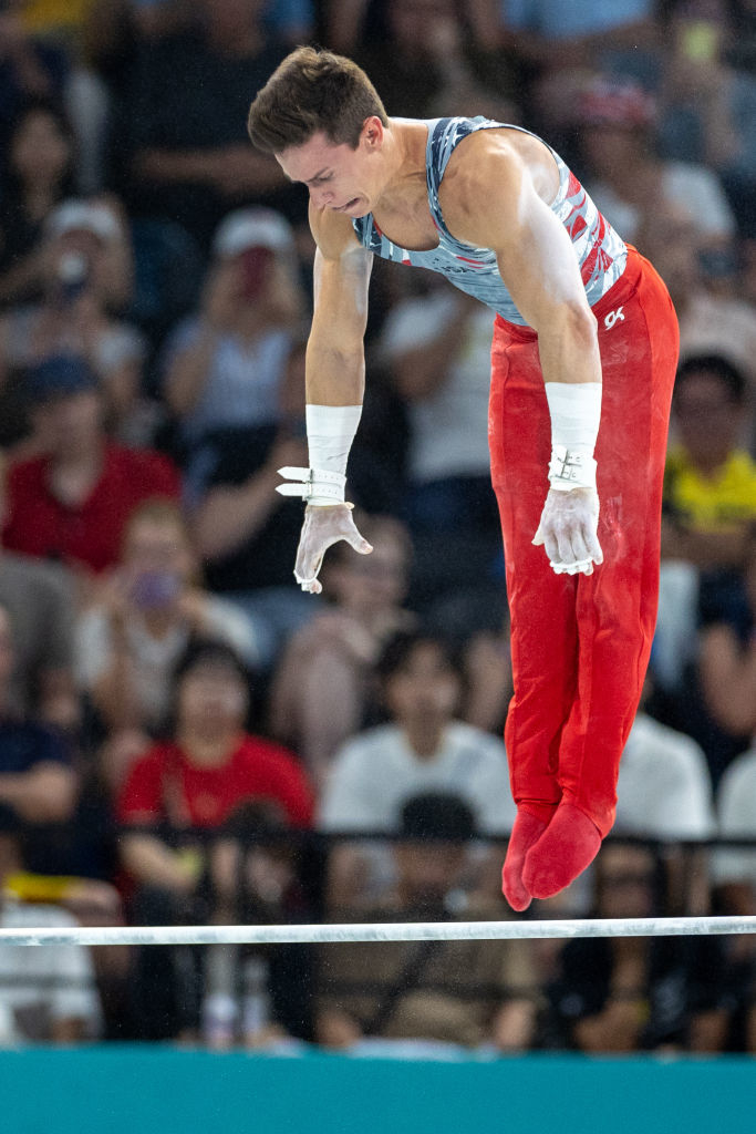 A gymnast performs a routine on the parallel bars at a competition, wearing red pants and a sleeveless top. The audience watches intently in the background