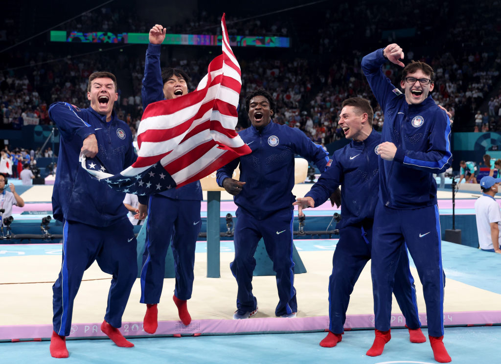 U.S. Olympic gymnast team wearing athletic tracksuits, holding and cheering with an American flag on the gymnastics floor
