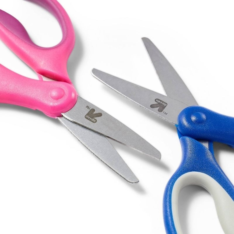 Two pairs of scissors with rounded tips, one with pink handles and the other with blue handles, are shown side by side