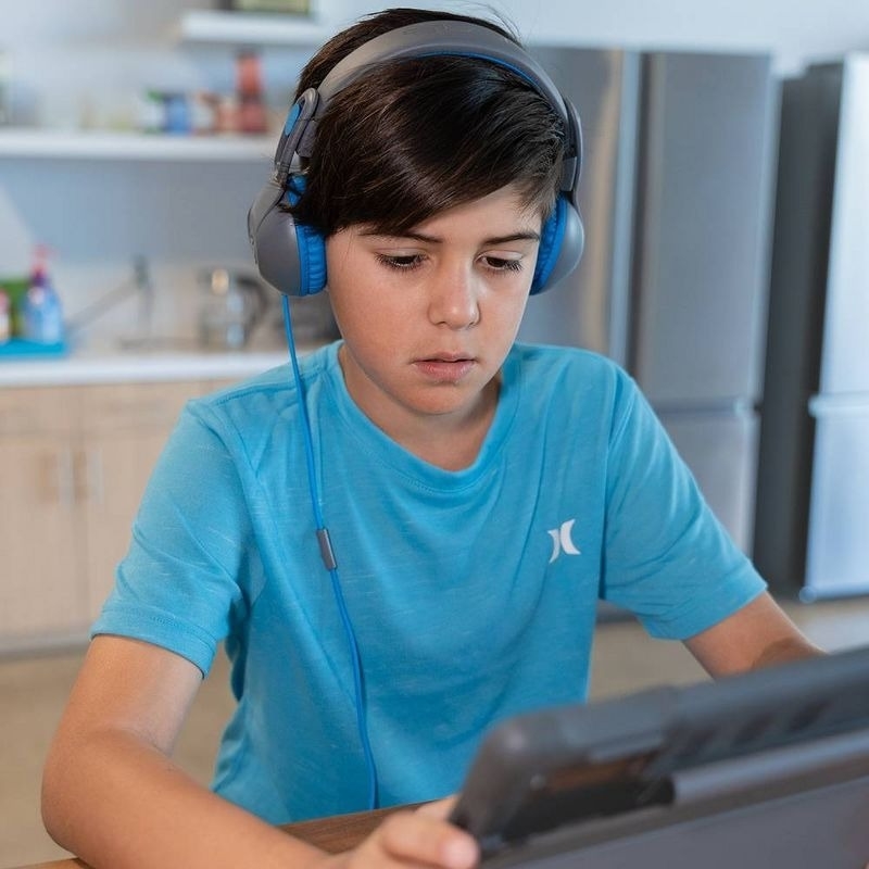 A boy wearing headphones looks intently at a tablet screen in a kitchen setting. He is wearing a turquoise shirt