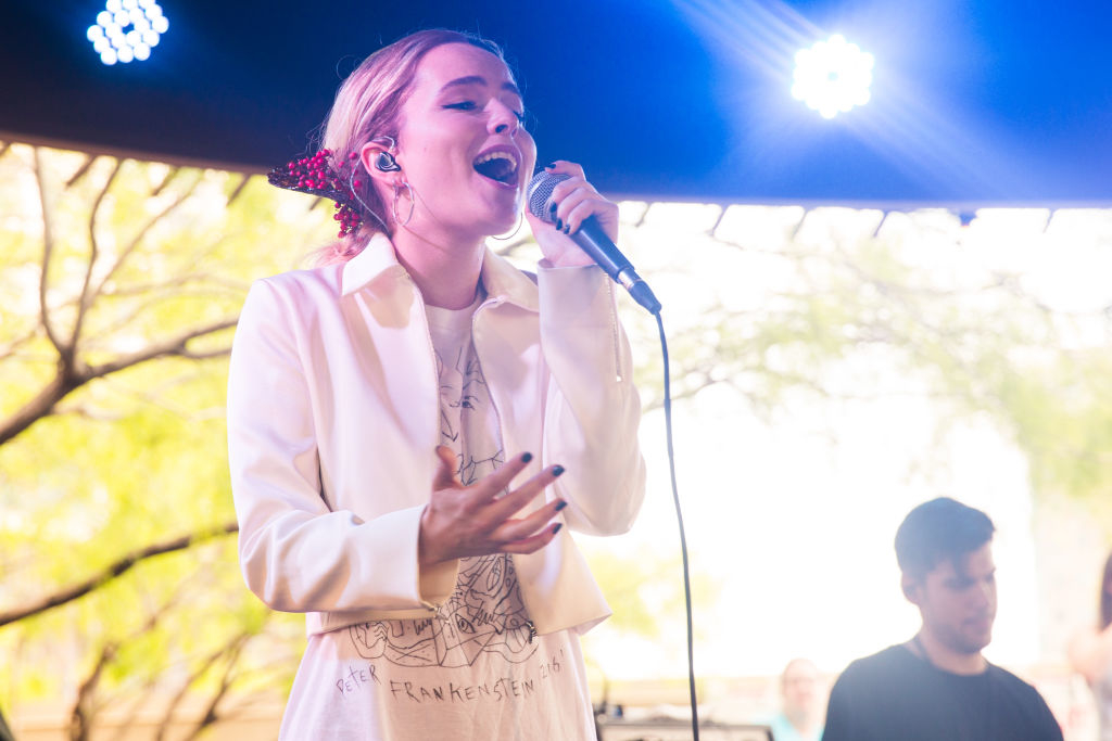Bridgit Mendler sings on stage in a casual outfit with a jacket. A musician is visible behind her