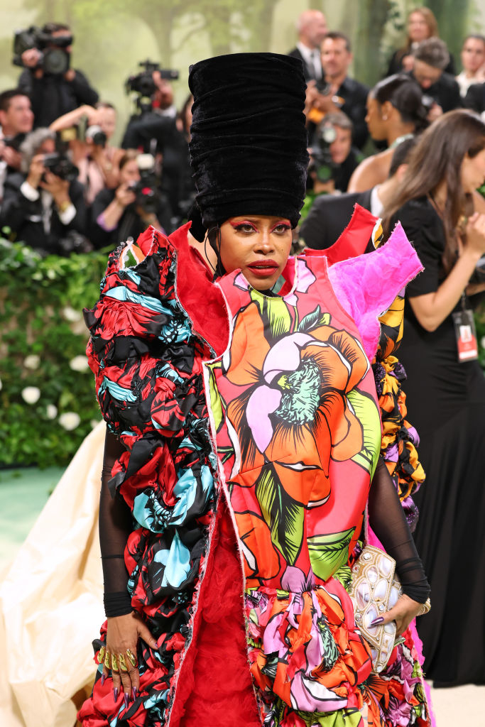 Erykah Badu on the red carpet wearing a vibrant, floral-themed avant-garde gown with a tall, dramatic headpiece