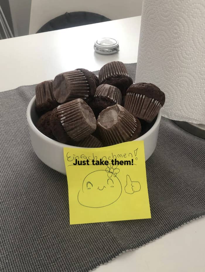 A bowl of chocolate muffins with a sticky note on the side displaying a drawing of a smiling face, thumbs up, and the text &quot;Just take them!&quot;
