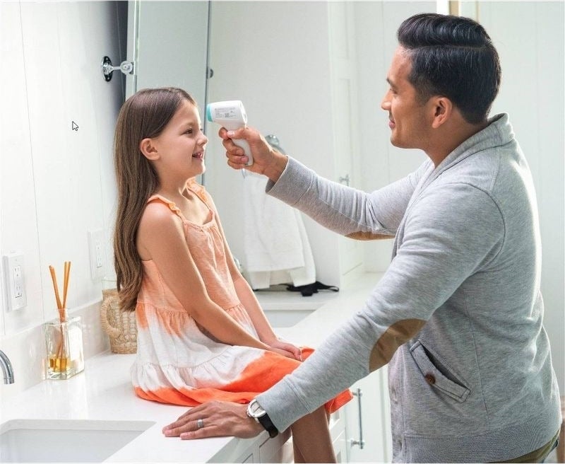 A man in a gray sweater uses a thermometer on a smiling girl in a sleeveless dress sitting on a bathroom counter