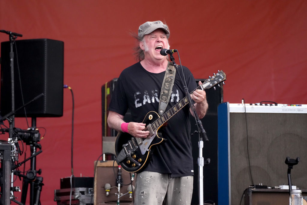 Neil Young performs on stage with an electric guitar, wearing a distressed t-shirt and jeans, and a cap