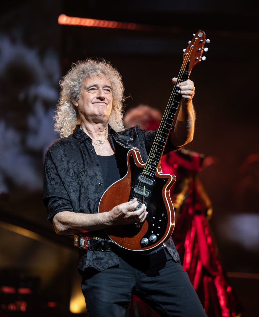 Brian May, wearing a patterned shirt, is playing an electric guitar on stage