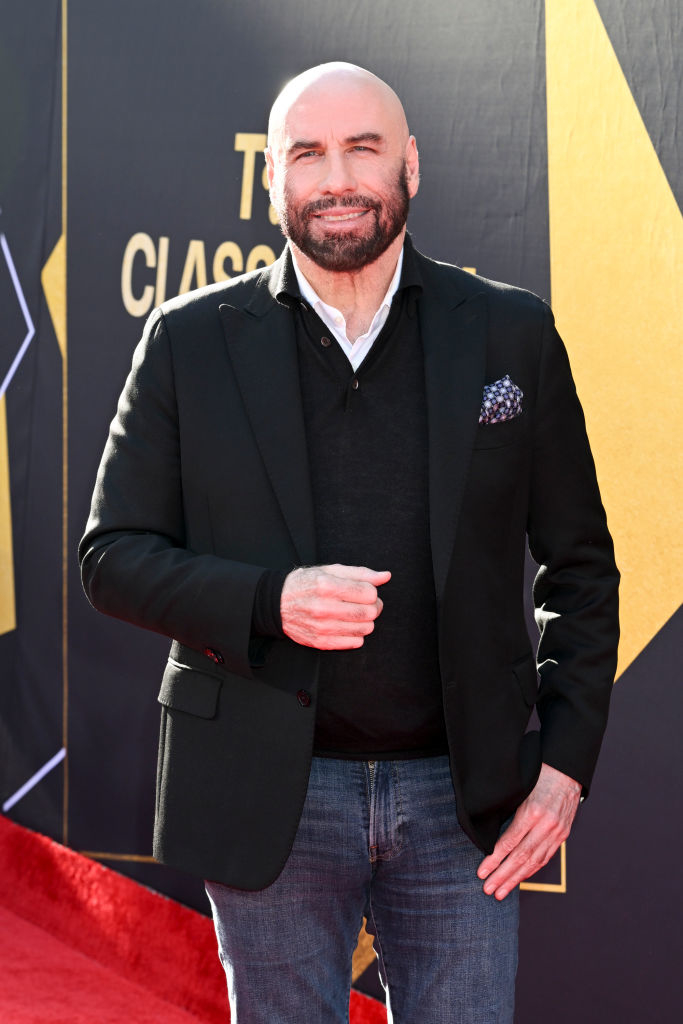 John Travolta poses on the red carpet wearing a jacket, sweater, collared shirt, and jeans at a celebrity event