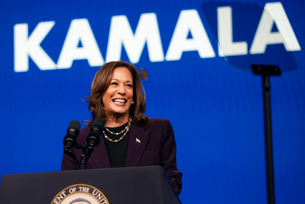Kamala Harris delivering a speech at a podium, with &quot;KAMALA&quot; displayed in large letters behind her. She is smiling and wearing a suit jacket