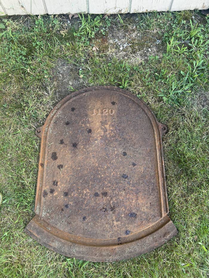 Flat rusty metal object in a tombstone-like shape labeled &quot;1120&quot; lying flat on grass