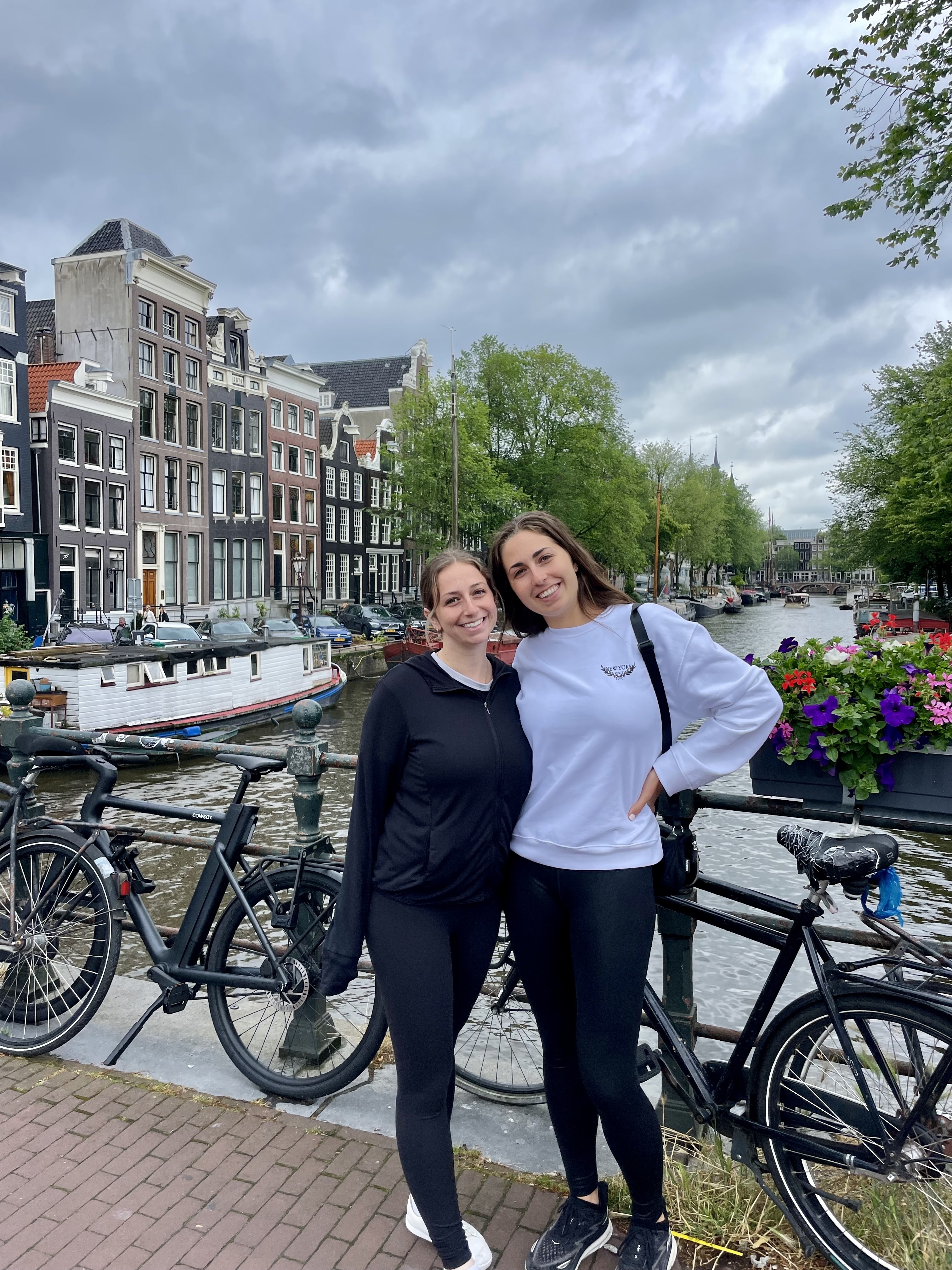 Two women in casual outfits pose by a canal in Amsterdam, with bikes and scenic architecture in the background. Names are not provided