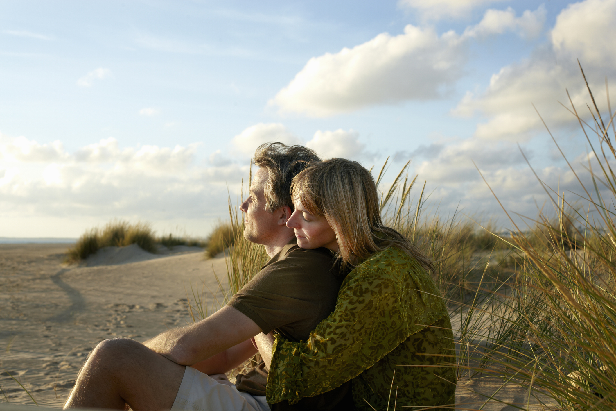 A man and a woman sit on a sandy beach with dunes and grasses, gazing at the sky. The woman is hugging the man from behind