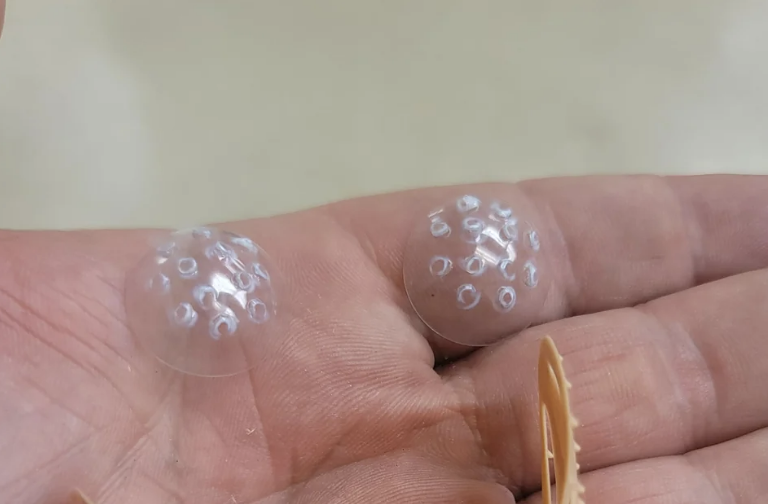 A hand holds two clear, dome-shaped objects with small circular patterns on them. A part of a beige object is shown in the background