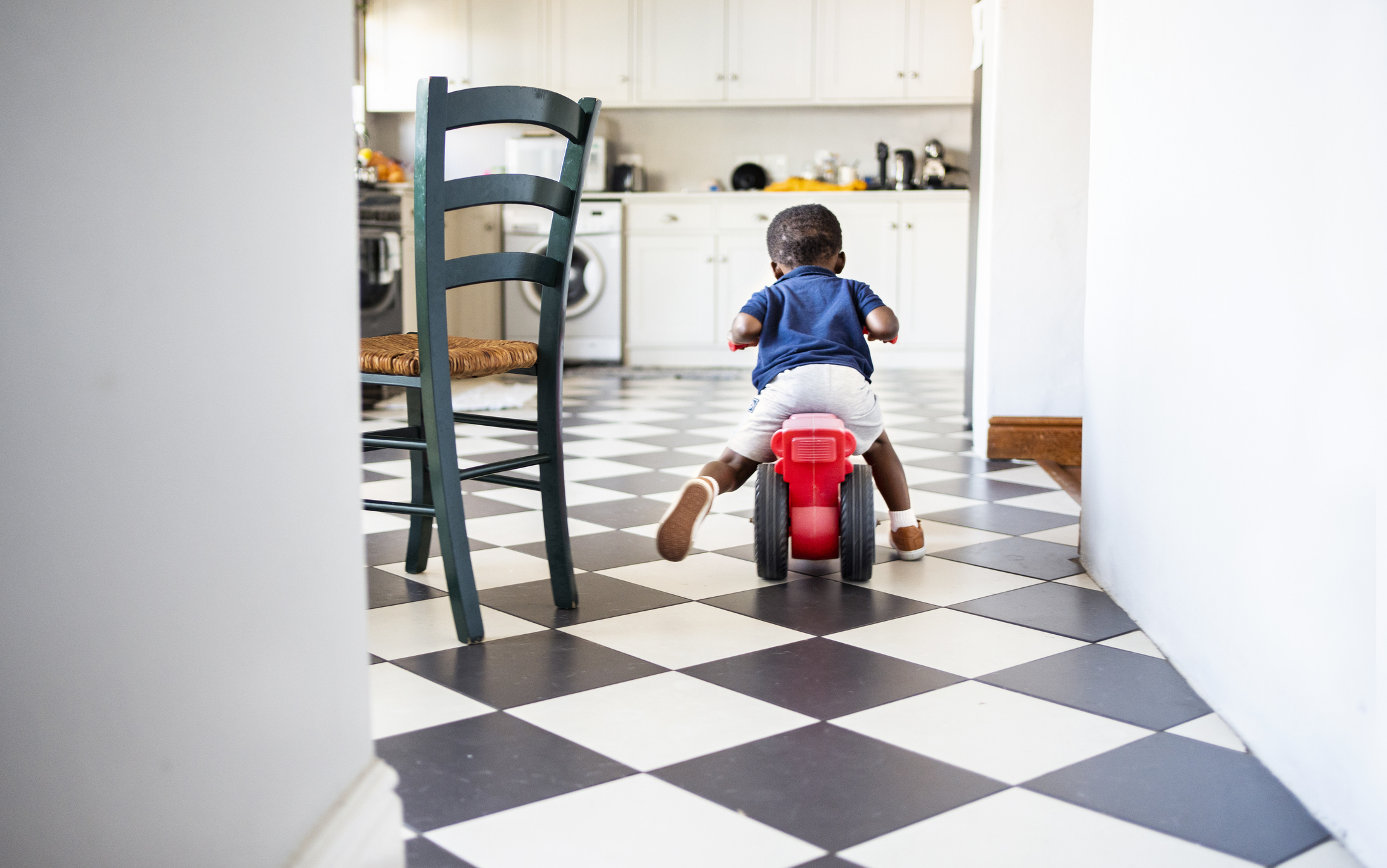 A small child rides a toy tricycle in a kitchen with checkered flooring and white cabinets