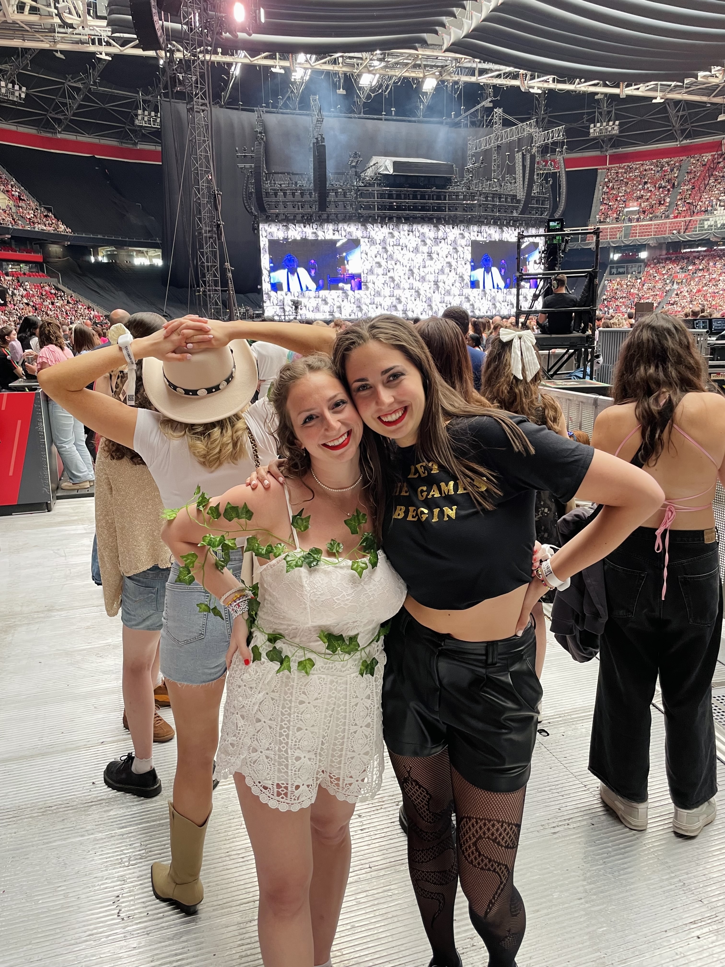 Two women smiling at a concert. One wears a white outfit with green leaf decorations, and the other wears black shorts, a black top, and fishnet stockings