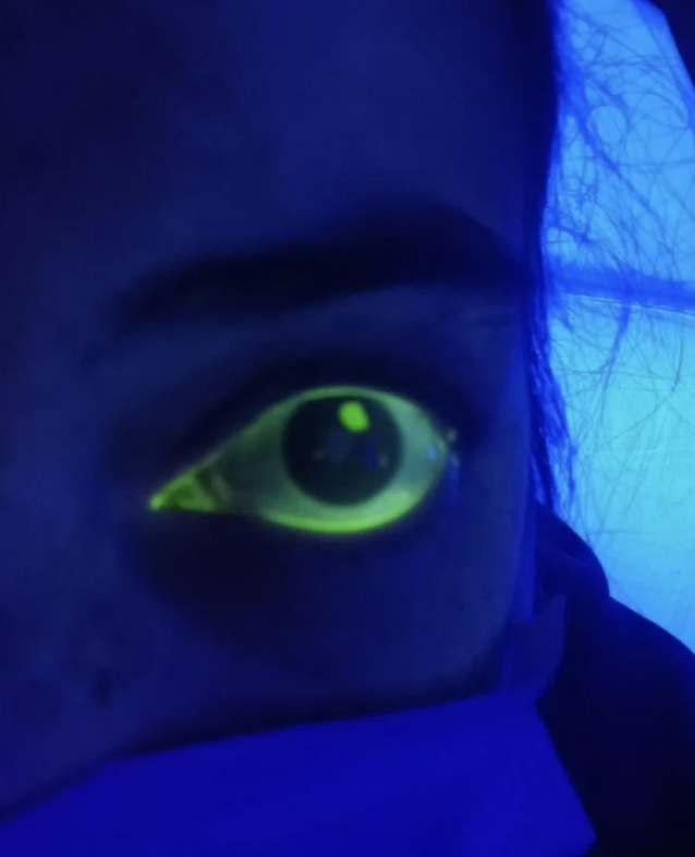 Close-up of a person's eye glowing brightly in blacklight with an artistic effect. Their face is partially covered by a cloth or garment. No identifiable persons