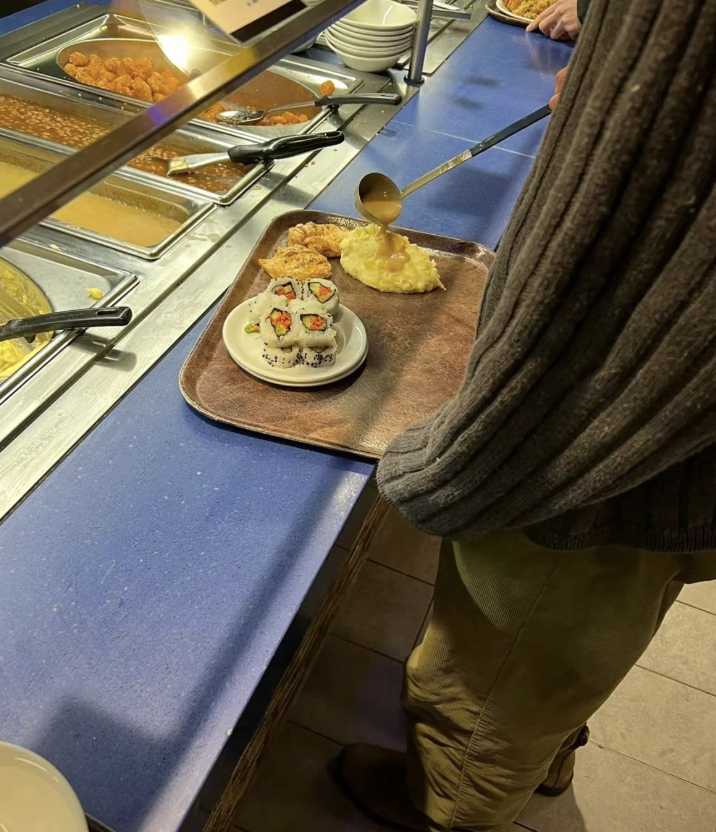 A person is serving food from a buffet, placing a scoop of mashed potatoes on a tray that has sushi and other items