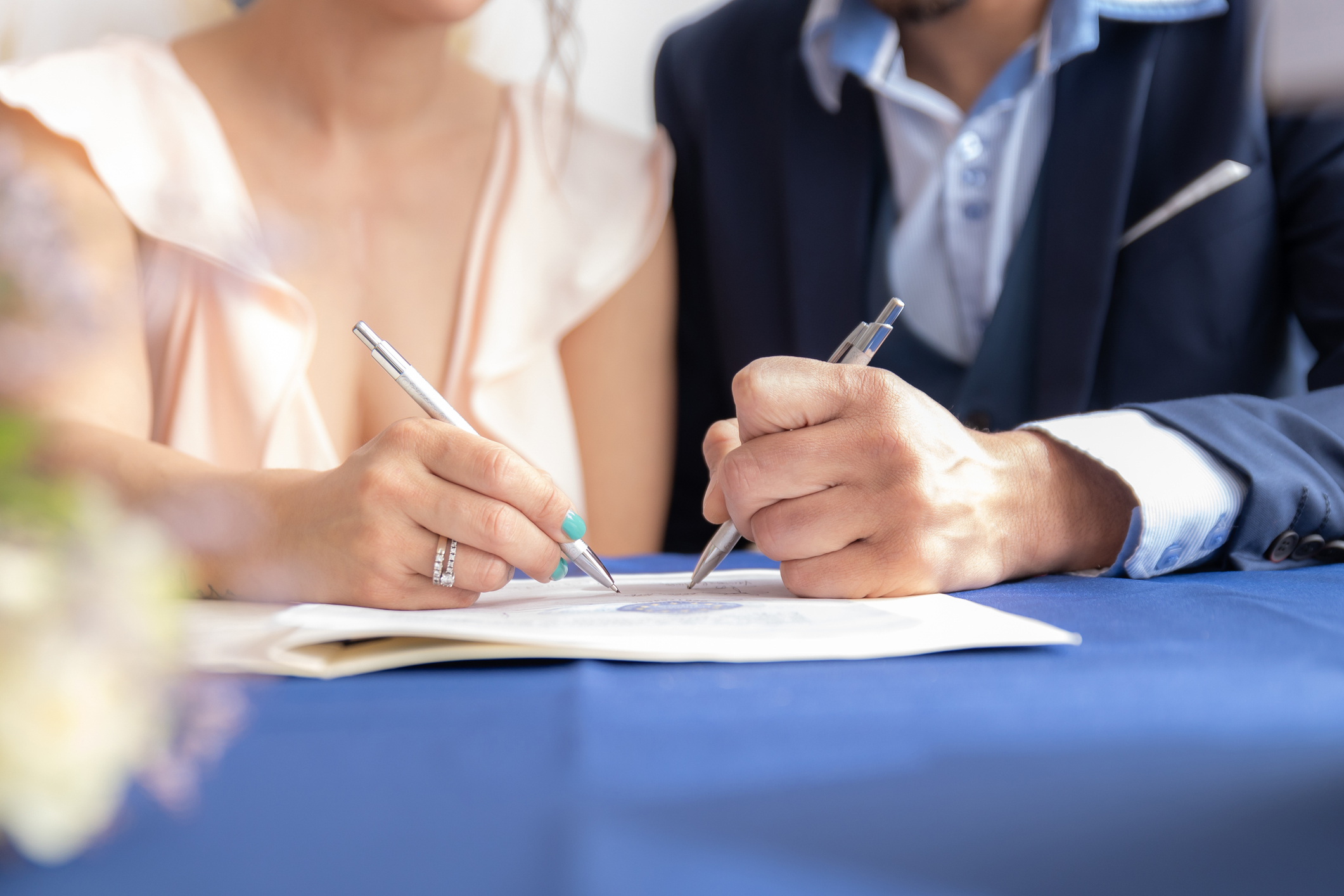 Two unidentified people signing a document, wearing formal attire. The person's nail polish and engagement ring are prominent
