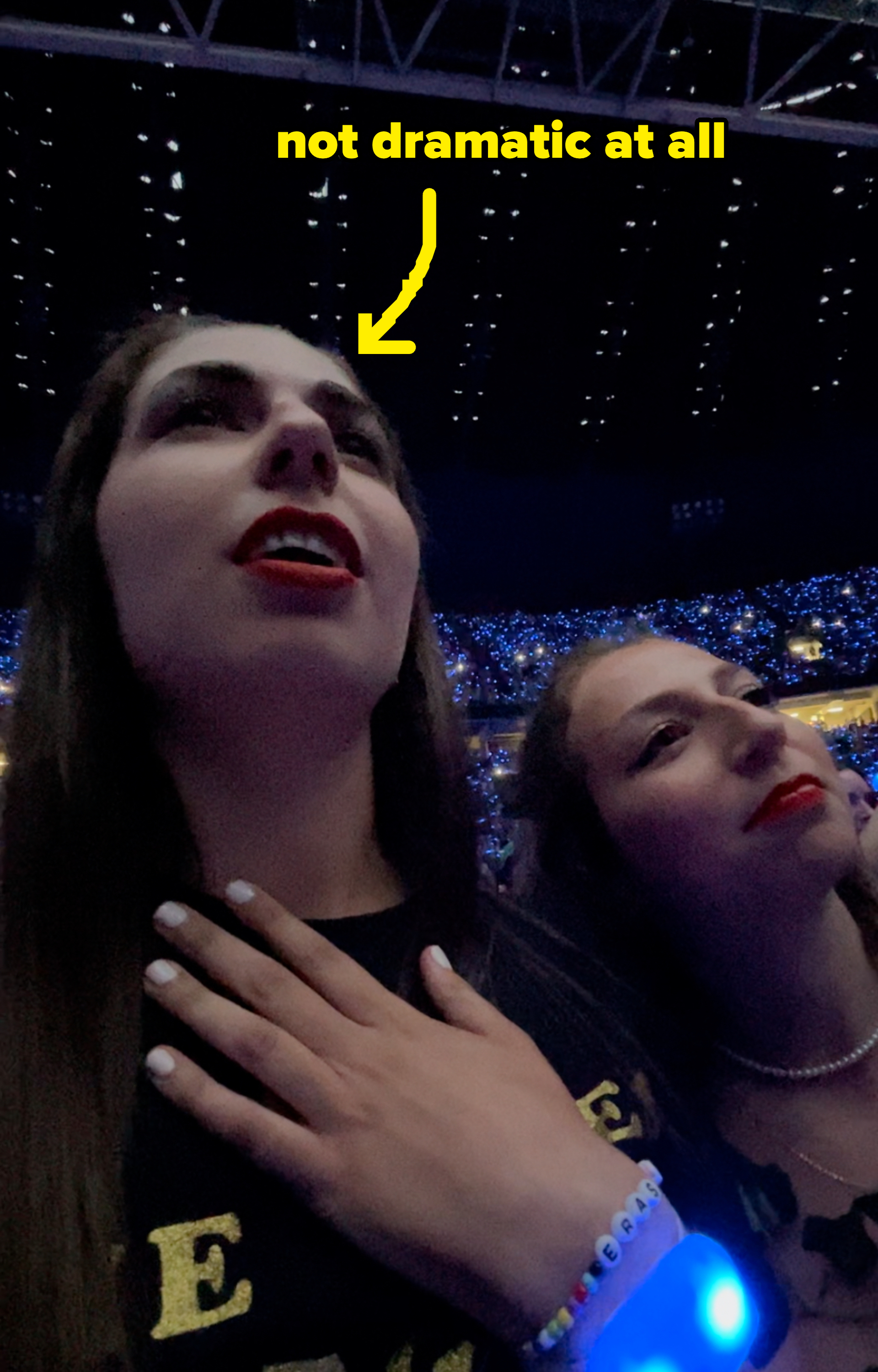 Two concertgoers sing along, one holding her hand to her chest, with a dome ceiling and a crowd with lit wristbands in the background