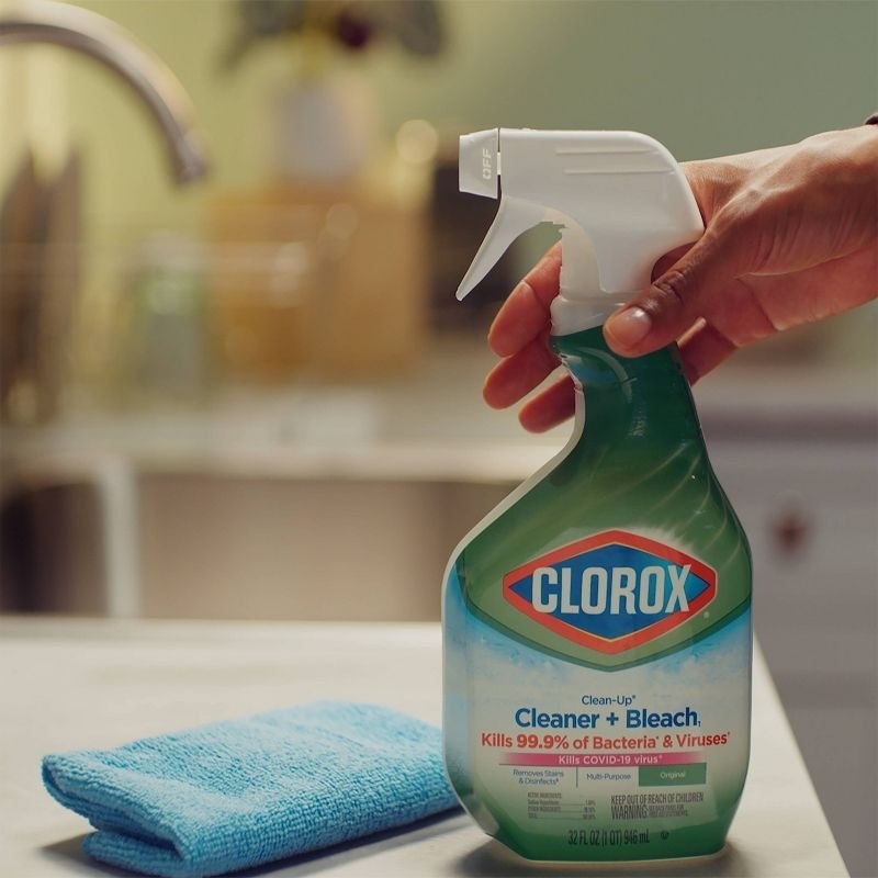 A hand holding a bottle of Clorox Cleaner + Bleach next to a blue cleaning cloth in a kitchen setting