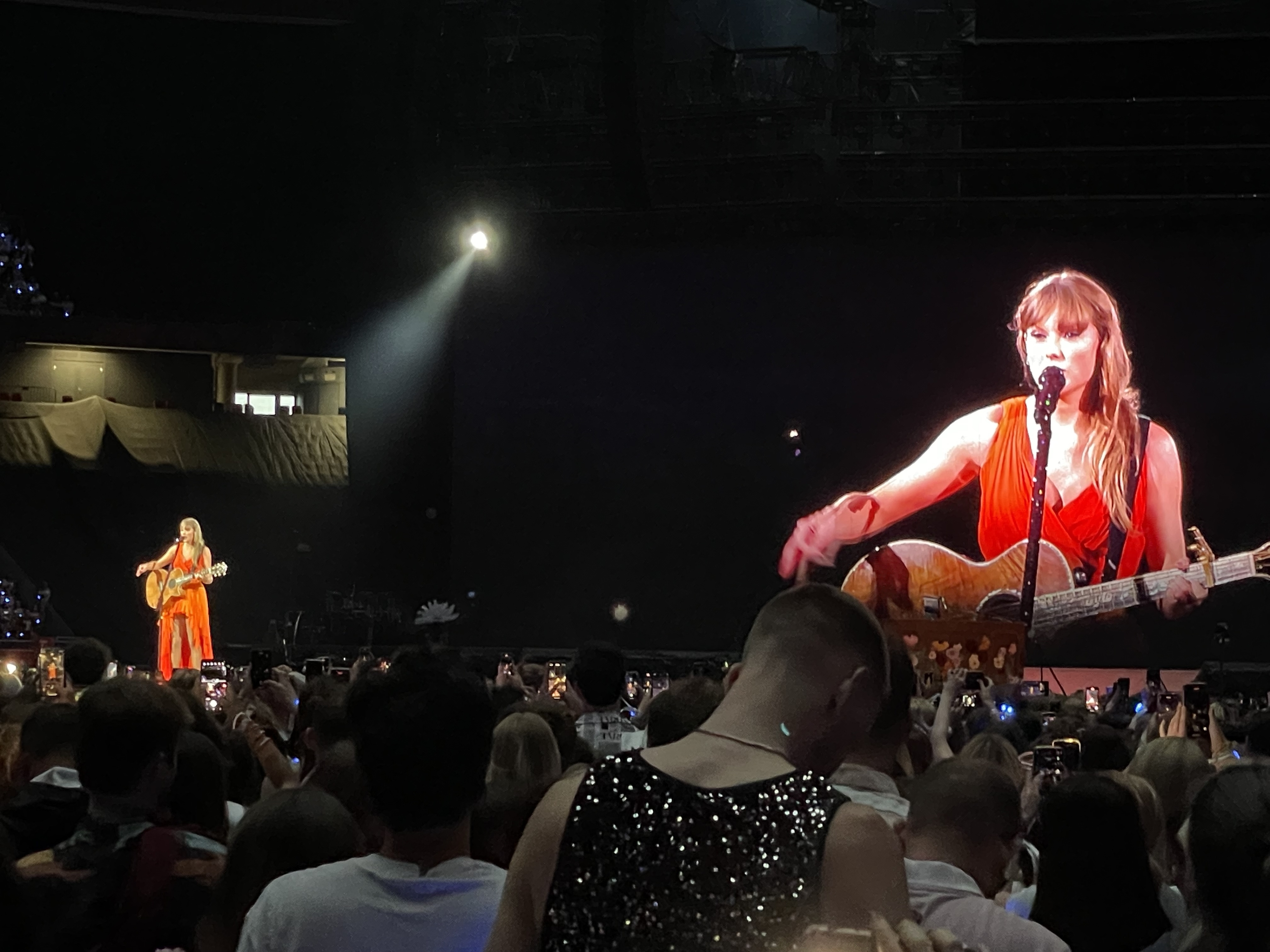 Taylor Swift performs onstage with a guitar at a concert; audience members watch and record the moment
