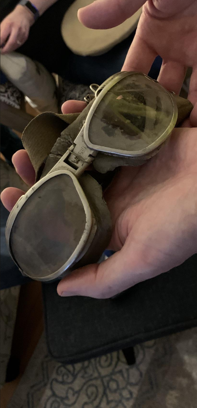 A pair of vintage, worn aviator goggles are being held in an individual’s hands. The lenses are discolored, and straps show signs of wear