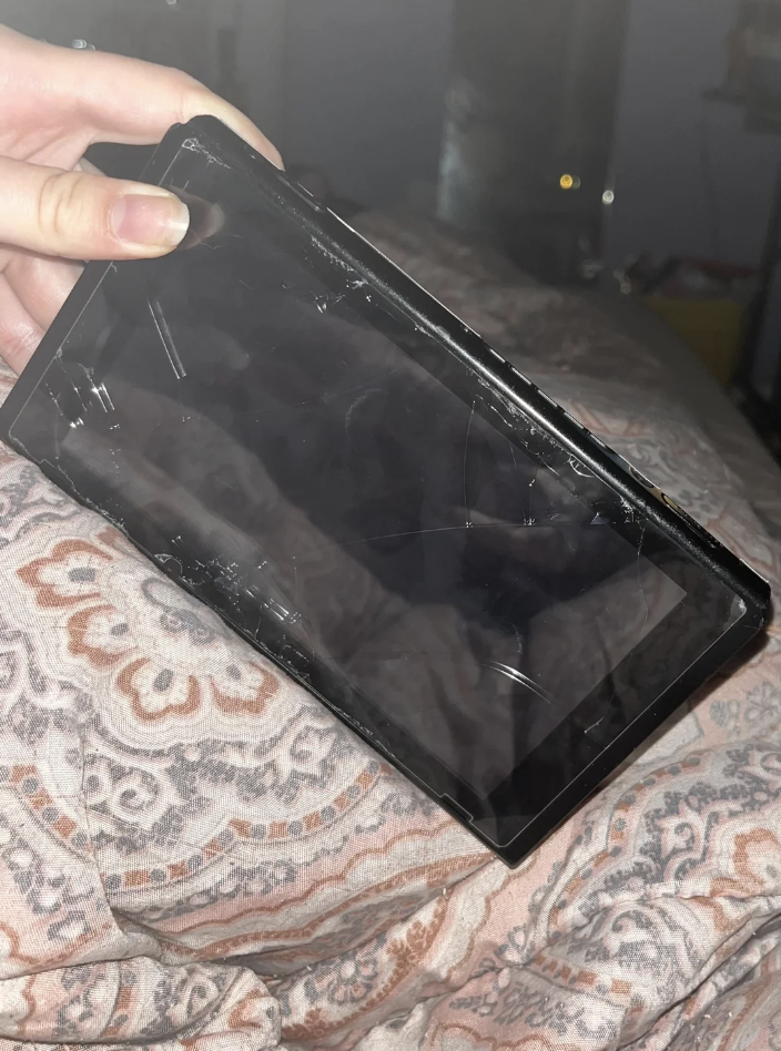 A hand holds a damaged tablet resting on a paisley-patterned fabric. The screen is severely cracked and partially detached from the frame