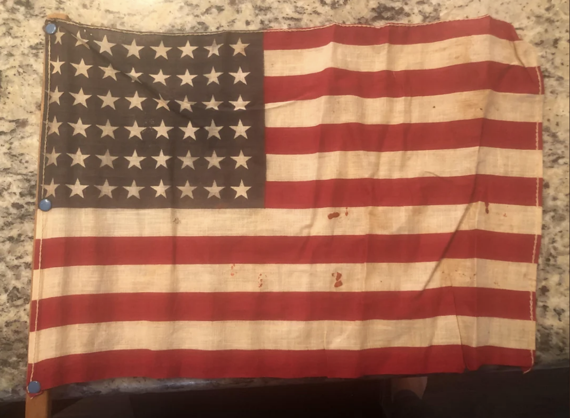 An aged United States flag with 48 stars, showing signs of wear and some stains, set against a speckled background