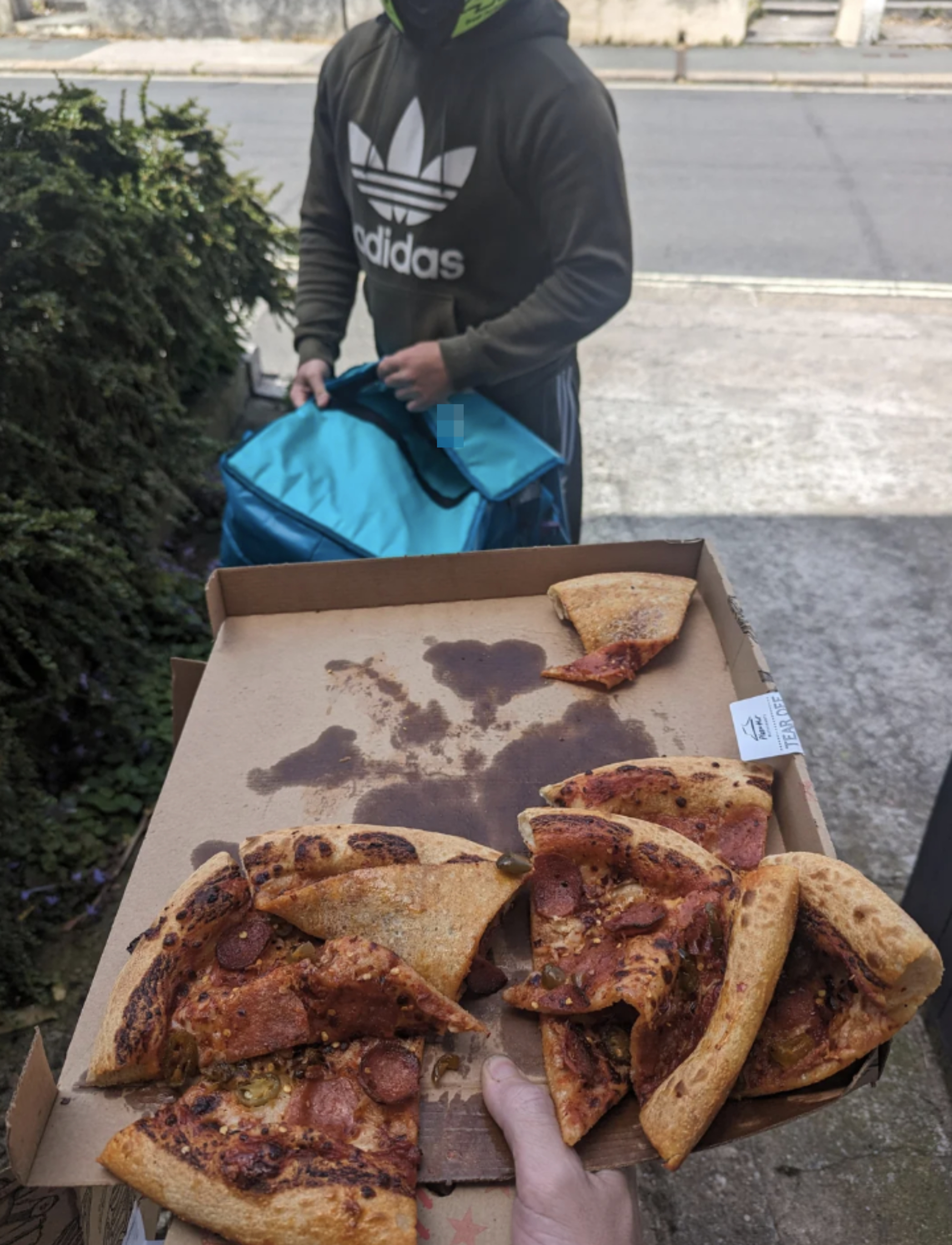 A person in an Adidas hoodie is delivering a pizza box with several pepperoni pizza slices to a home. The person is holding an insulated pizza delivery bag