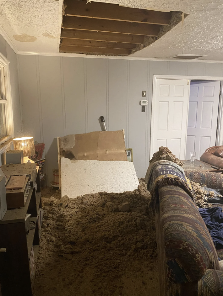 A living room with a collapsed ceiling scattering debris and insulation onto the floor and furniture, including a patterned couch. A lamp and bookshelf are visible on the left