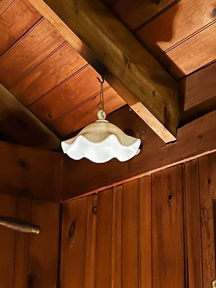 A rustic wooden ceiling with a vintage scalloped ceramic ruffled item hanging from a hook. The light fixture is white with a curved edge design