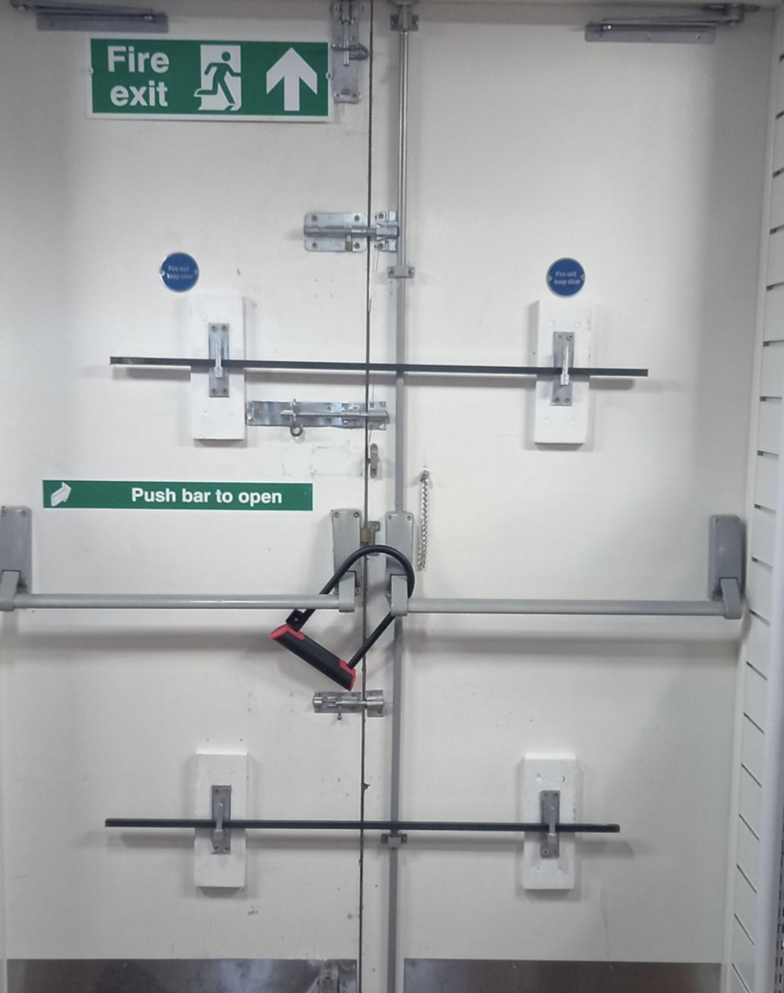 Fire exit door heavily barricaded with multiple locks and poles; a chain and padlock also secure the door. Sign above reads &quot;Fire exit&quot; with an arrow and exit symbol