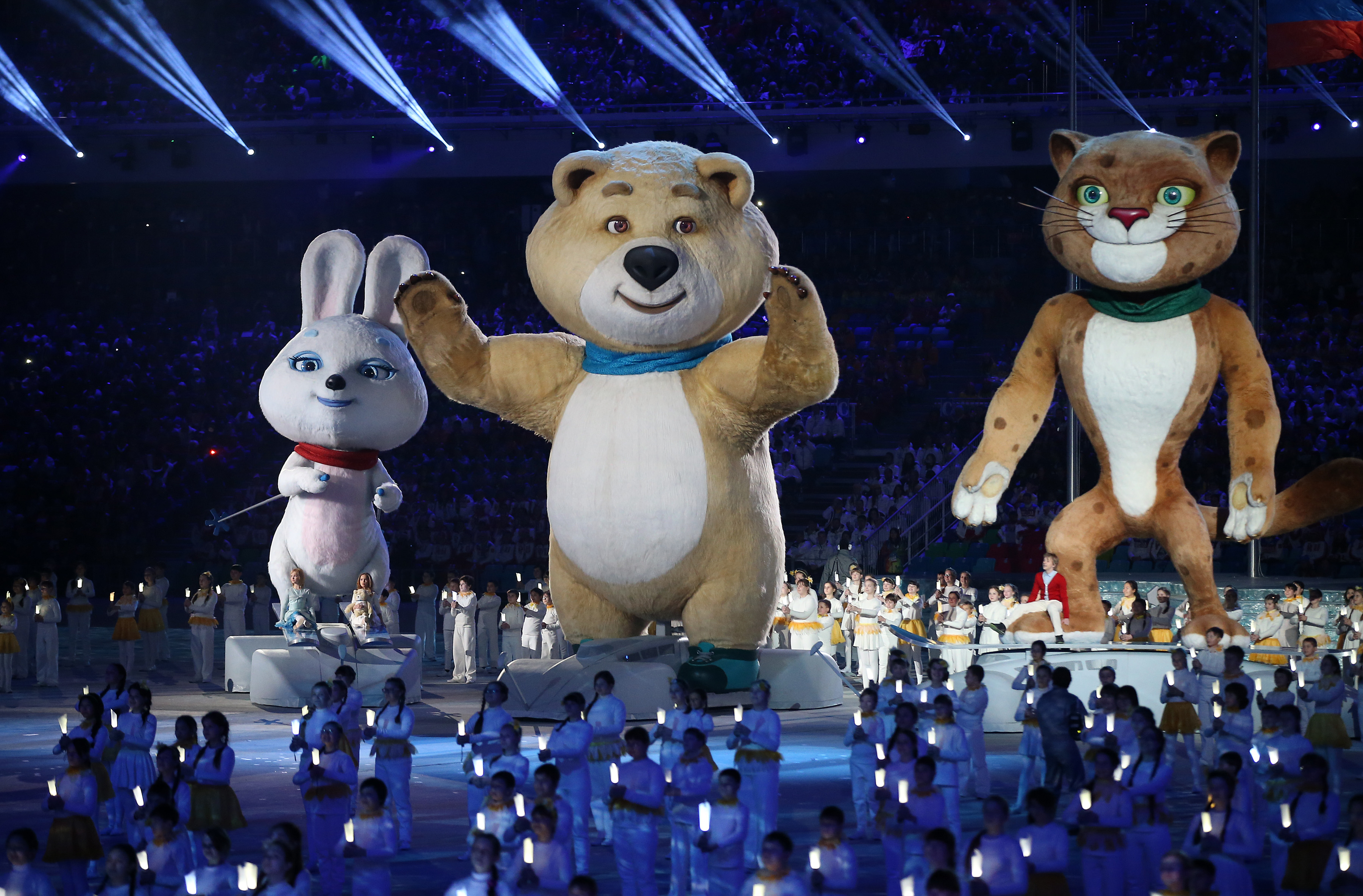 Three large mascots—a white rabbit, a beige bear, and a brown leopard—stand on stage at an event, surrounded by performers in white outfits holding lights