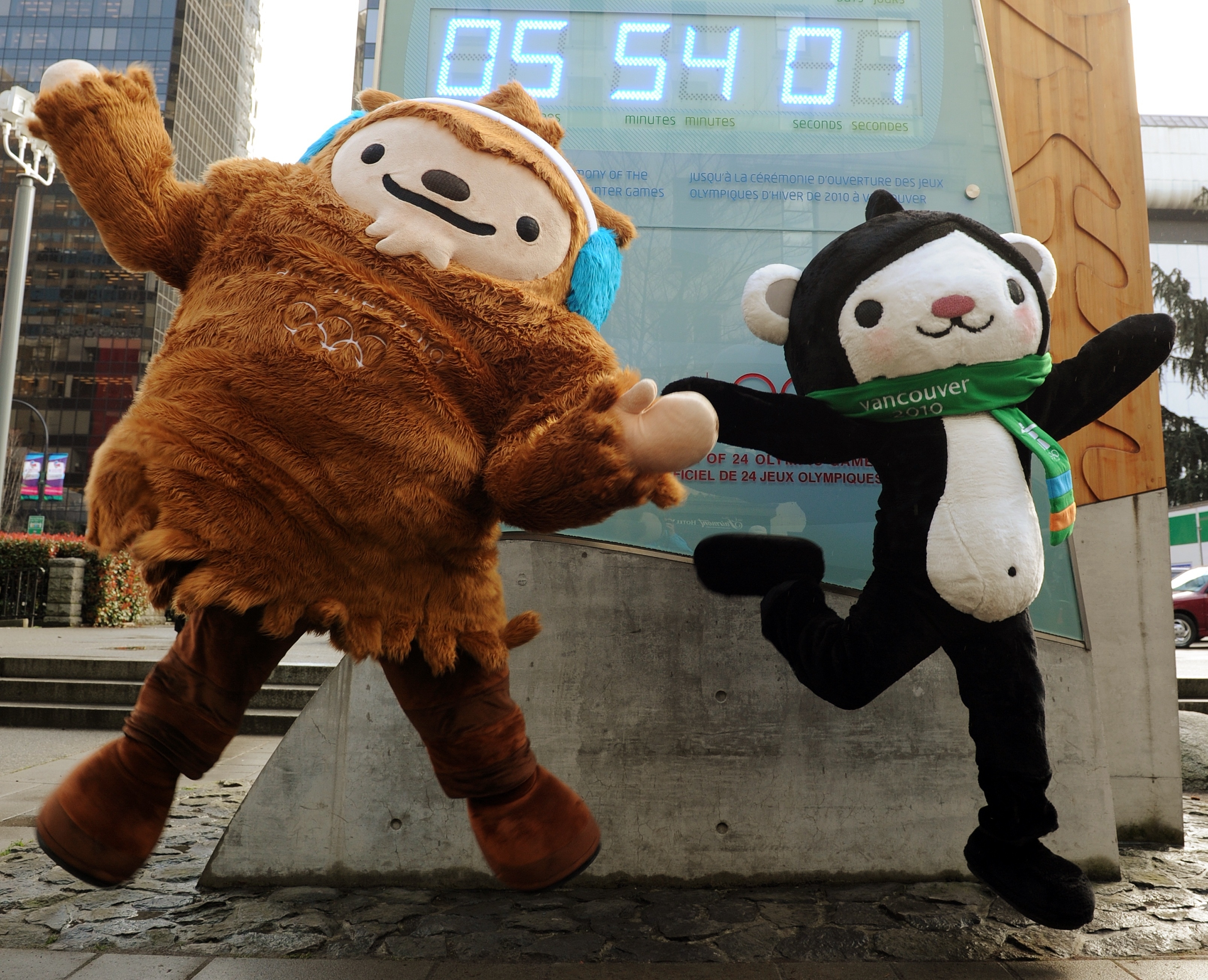 Sumi and Quatchi, mascots of the 2010 Winter Olympics in Vancouver, leap joyfully in front of a digital countdown timer. Sumi is dressed as a bear with wings, and Quatchi is a sasquatch in earmuffs