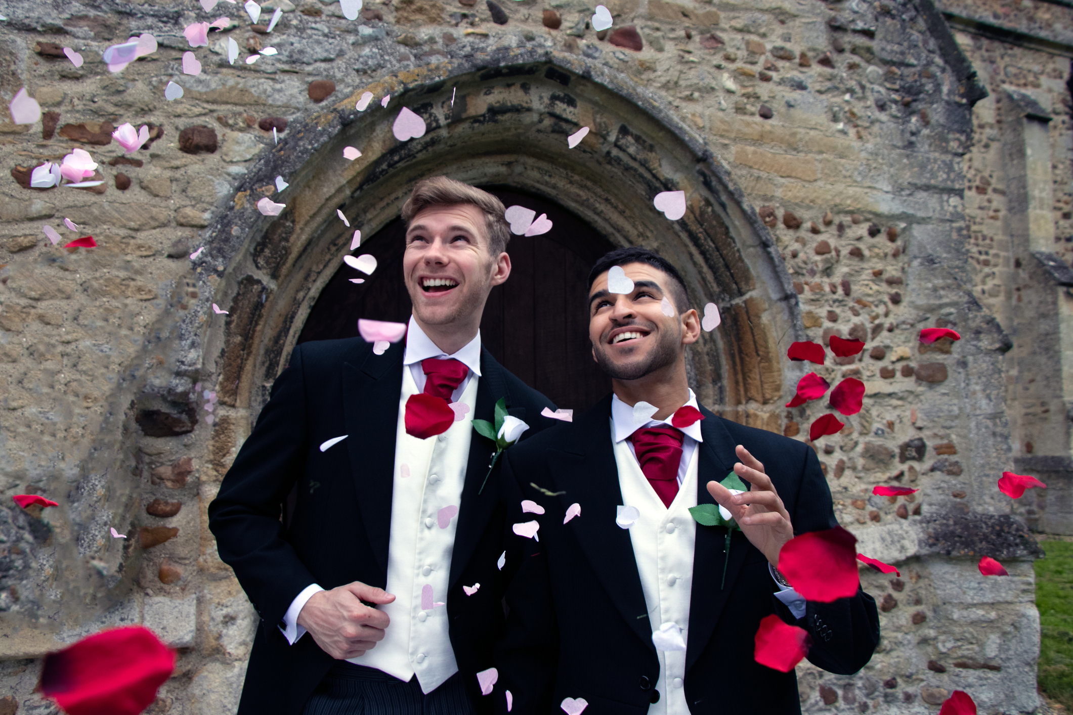 Two men in suits, smiling as rose petals are thrown, standing in front of a stone building, celebrating a wedding