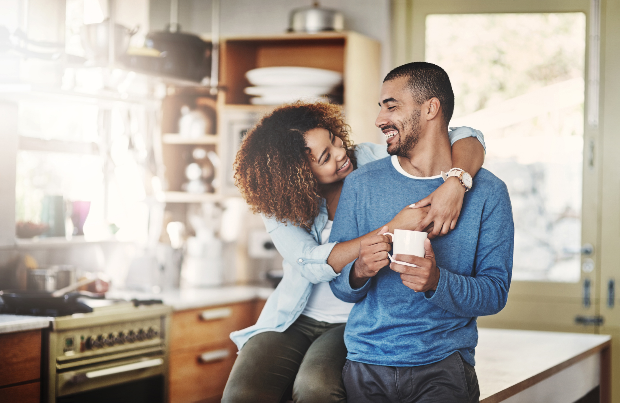 A woman sits on a kitchen counter, affectionately hugging a man who holds a mug and smiles at her. Both are casually dressed in a cozy kitchen setting