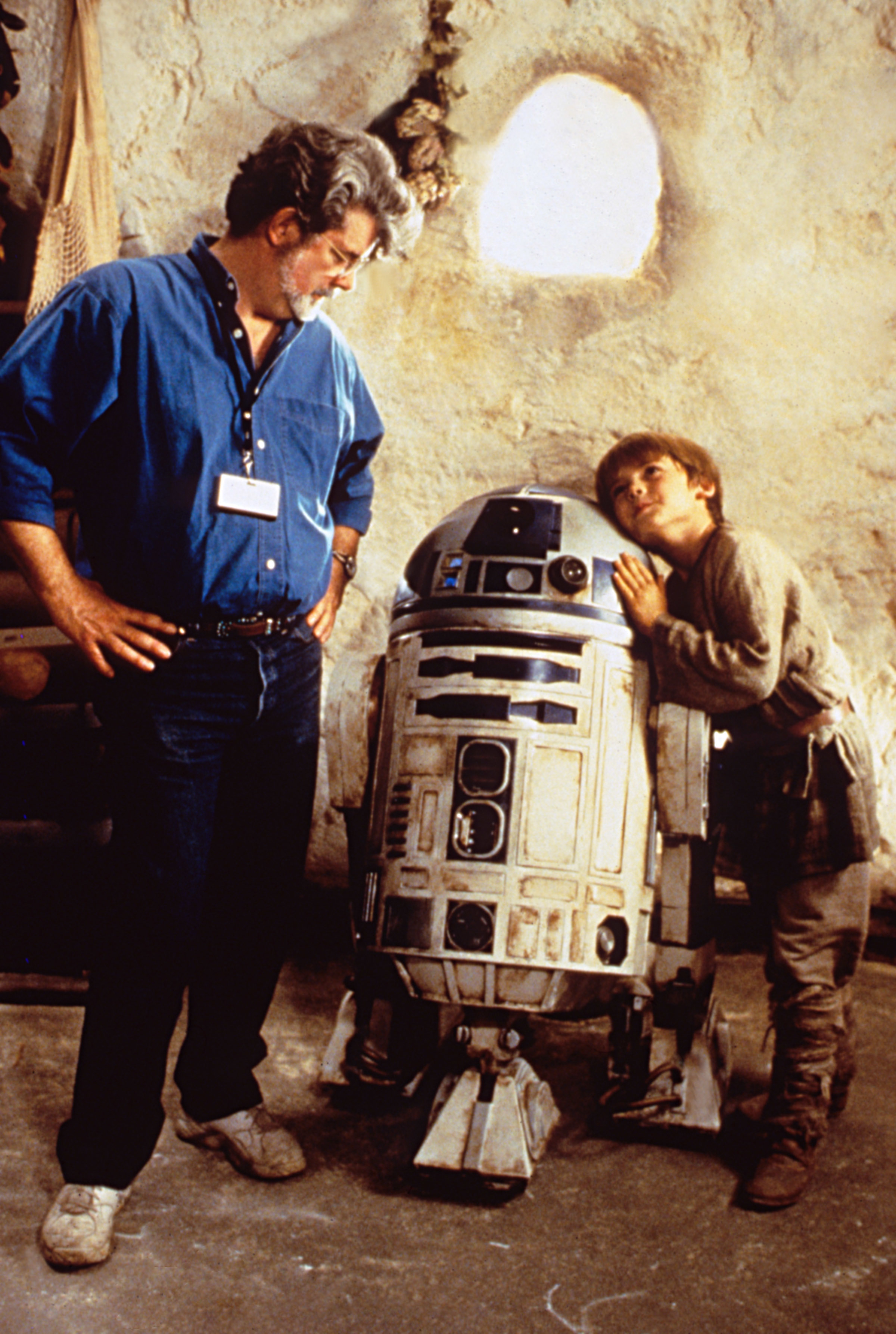 George Lucas standing next to R2-D2 with a young Jake Lloyd leaning on the droid, in a set resembling Tatooine